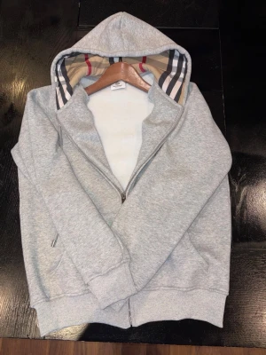 Grå zip hoodie från Burberry - Snygg grå zip hoodie från Burberry med klassiskt rutigt foder i huvan och dragkedja med Burberry-detalj. Tillverkad i mjukt bomullsmaterial och har ribbade muddar vid ärmslut och nederkant. Perfekt för en chill och stilren look. Storlek M men passar S. Pris kan diskuteras!