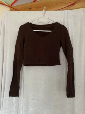 Brun croppad långärmad topp - Mörkbrun croppad topp med lång ärm och v-ringning. Enkel och stilren design som passar perfekt till jeans eller kjol. Mjuk och stretchig känsla, troligtvis i bomull eller polyesterblandning.