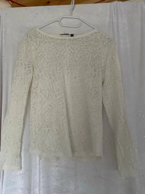 Vit spetsblus med rosor från H&M - Vit långärmad blus från H&M i transparent spets med rosor som mönster. Blusen har en rak passform och rund halsringning, perfekt för att skapa en romantisk look. Materialet är lätt och luftigt, vilket gör den superbekväm att bära.