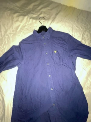 Marinblå skjorta från Lyle & Scott - Snygg marinblå skjorta från Lyle & Scott med klassisk krage och knappstängning framtill. Skjortan har lång ärm och den ikoniska gula loggan broderad på bröstet. Perfekt för dig som gillar stilrena plagg med en sportig touch.