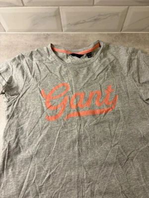 Grå Gant t-shirt med rosa tryck - Snygg grå t-shirt från Gant med stort rosa logotyptryck på bröstet. Klassisk rund halsringning och korta ärmar. Tillverkad i mjuk bomull som är skön att ha på sig. Perfekt för en avslappnad och trendig stil. Den kommer såklar strykas innan den skickas iväg