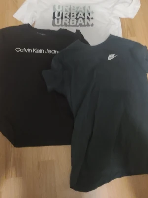 T-shirtpaket Nike, Calvin Klein & Urban - Tre snygga t-shirts: en svart Nike med vit logga, en svart Calvin Klein Jeans med vit text och en vit Urban med grått tryck. Alla har klassisk passform och rund hals. Perfekta för en stilren och sportig look. Använda säljer för 199kr pris inte hugget i sten!