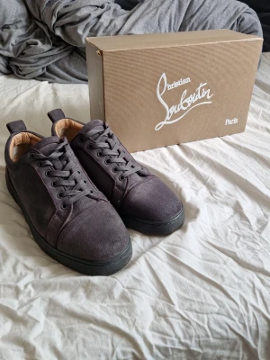 Christian Louboutin gråa sneakers - Snygga svarta sneakers från Christian Louboutin med klassisk röd sula och diskret logga undertill. Skorna har en låg profil, rund tå och är tillverkade i mocka med gråa skosnören. Perfekta för dig som vill ha en stilren och exklusiv look.