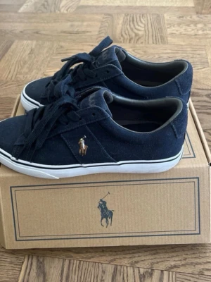 Mörkblå Polo Ralph Lauren sneakers - Snygga mörkblå sneakers från Polo Ralph Lauren med klassisk logga på sidan. Skorna har lågt skaft, vita sulor och är tillverkade i mocka med textilfoder. Perfekta för dig som gillar stilrena och tidlösa sneakers. Skorna är i storlek 39 och passar perfekt för båda män och kvinnor. Aldrig använda och helt nya, kommer med lådan också. Köpta för 1500 så väldigt bra pris för helt nya skor.  Kontakta om ni har några frågor eller bud osv. 