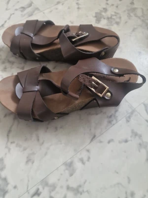 Bruna kilklackade sandaler More More - Snygga bruna sandaler från More More med kilklack och breda remmar i läderimitation. Spänne i guldton och öppen tå ger en trendig look. Perfekta för dig som gillar en stilren och bekväm sko med lite höjd. Priset kan diskuteras🫶🏼