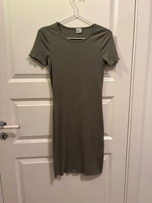 Grön ribbad klänning från Gina Tricot - En enkel och stilren grön ribbad klänning från Gina Tricot. Klänningen har korta ärmar, rund halsringning och en kroppsnära passform som slutar strax under knäna. Perfekt för en clean och avslappnad look. Jag har tidigare haft katter så katthår kan förekomma men kläderna är alltid tvättade innan de skickas. 