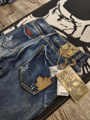 Dsquared2 blå slitna jeans - Säljer ett par blå jeans från Dsquared2 med slitningar, kontrastsömmar och detaljer som röd patch och guldfärgad lönnlöv-applikation. Jeansen har klassisk femficksmodell och unika Dsquared2-taggar bak.