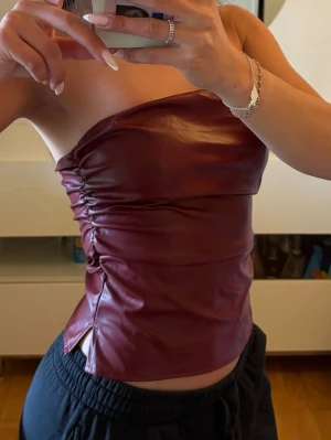 Vinröd tubtopp i skinnimitation - Snygg vinröd tubtopp i skinnimitation med rynkade detaljer på sidan och liten slits nedtill. Toppen är axelbandslös och har en tight passform som framhäver figuren. Perfekt för dig som vill sticka ut med en trendig look.