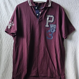 Vinröd pikétröja från U.S. Polo Assn. - Vinröd pikétröja med korta ärmar från U.S. Polo Assn. Tröjan har broderade detaljer och tryck på bröstet samt ärmen, klassisk krage med knappar och kontrastfärgade ränder på kragen. Perfekt för en sportig och avslappnad stil.