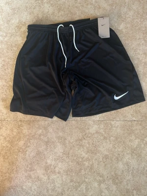 Svarta Nike shorts med snörning - Svarta sportiga shorts från Nike med vit snörning i midjan och klassisk vit Nike-logga på ena benet. Tillverkade i ett lätt och svalt polyestermaterial som passar perfekt för träning eller chill. Elastisk midja för skön passform.