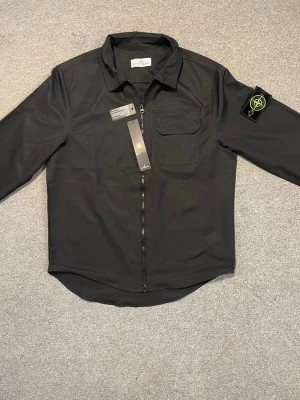 Svart overshirt från Stone Island - Snygg svart overshirt från Stone Island med dragkedja framtill, klassisk krage och bröstficka. Ikonisk logotyp-patch på vänster ärm. Tillverkad i ett slätt och lätt material som ger en clean look. Perfekt för lager-på-lager-stil.