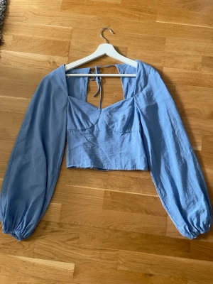 Ljusblå croppad blus med puffärm - Trendig ljusblå blus med croppad passform och långa puffärmar. Toppen har en snygg v-ringning, öppen rygg med knytband och smock baktill för extra komfort.