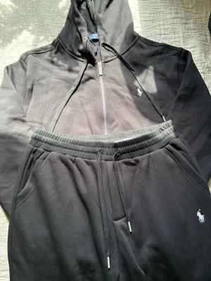Svart hoodie från Polo Ralph Lauren - Svart hoodie med dragkedja från Polo Ralph Lauren. Klassisk design med huva och dragsnören, samt den ikoniska vita logotypen broderad på bröstet. Tillverkad i mjukt bomullsmaterial och har fickor framtill. Perfekt för en avslappnad och stilren look.