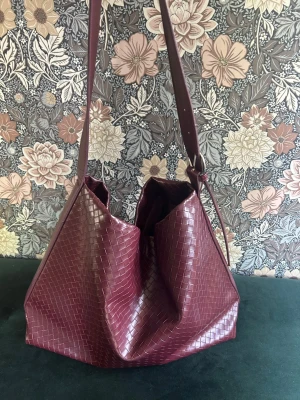 Vinröd axelväska i flätad look - Snygg vinröd axelväska med flätad läderimitation och bred axelrem. Väskan har en rymlig och mjuk form, perfekt för dig som gillar en stilren men ändå trendig accessoar. Passar till många olika outfits och ger en cool touch till din stil.Är i mycket bra skick!!har inga defekter men har används några gånger så inte helt ny men ser ut som den är ny!!