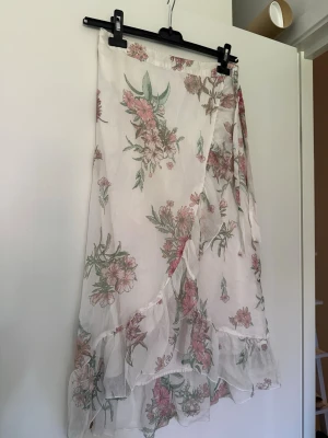Blommig omlottkjol med volang - Söt omlottkjol i vitt med blommigt mönster i rosa och grönt. Kjolen har volangkant och knytband i midjan, vilket ger en romantisk och luftig känsla. Perfekt för varma dagar och somriga outfits.