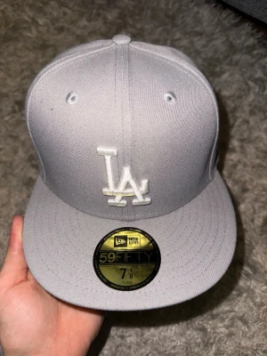 Grå LA New Era 59Fifty keps - Snygg grå snapback från New Era med LA-logga broderad framtill i vitt. Klassisk 59Fifty-modell med rak skärm och broderade ventilationshål. Perfekt för dig som gillar streetwear och vill ha en stilren look.