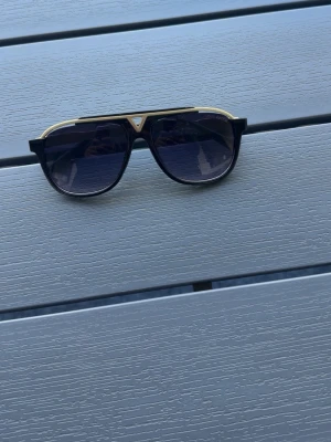 Glasögon - Stilrena svarta solglasögon med rektangulära bågar och detaljer i guld på skalmarna. Perfekt accessoar för att lyfta din look med en lyxig touch. Klassisk design som passar till många stilar.