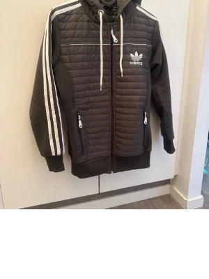 Svart Adidas hoodie med dragkedja - Snygg grön hoodie från Adidas med quiltade paneler framtill, vita ränder längs ärmarna och vit Adidas-logga på bröstet. Utrustad med huva och dragsko samt två fickor med dragkedja. Perfekt för en sportig och avslappnad stil.
