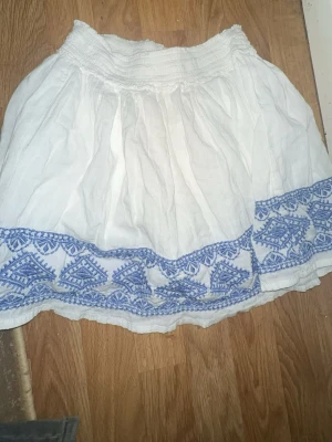 Vit kortkjol med blå brodyr från L.O.G.G. - Söt vit kortkjol med blå broderade detaljer längs nederkanten. Kjolen har resår i midjan och är gjord i ett lätt och luftigt bomullstyg, perfekt för varma dagar. Har aldrig använt den