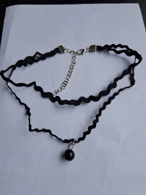 Svart chokerhalsband med pärla - Dubbelvågat svart chokerhalsband med en liten svart pärlberlock. Halsbandet har justerbart silverfärgat spänne och kedja. Perfekt accessoar för en edgy och trendig look.