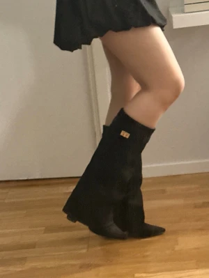 Svarta stövlar med klack och gulddetalj - Snygga svarta stövlar med spetsig tå och klack. Tillverkade i mockaimitation och dekorerade med en liten guldfärgad logga vid skaftet. Dragkedja på insidan för enkel på- och avtagning. Perfekta till kjol eller klänning för en trendig look. Det är ca 10 cm klack. Defekt på ena stöveln sulan sitter lite konstigt, jag tycker inte de känns så mycket och det kanske går att fixa men för kännedom så sitter ena sulan lite konstigt. Jag tror dom är i storlek 39 men kan också vara 38.