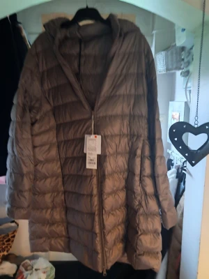 Ljusbrun lång dunjacka med huva - Snygg ljusbrun dunjacka från Noisy May med lång passform och huva. Jackan har dragkedja framtill och quiltad design som ger en modern look. Perfekt för kalla vinterdagar och enkel att matcha med olika outfits.