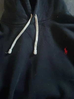 Svart hoodie från Polo Ralph Lauren. Tar bud - Svart hoodie från Polo Ralph Lauren med klassisk röd broderad logga på bröstet. Tröjan har huva med vita dragsnören och ribbade muddar vid ärmslut och nederkant. Perfekt för en chill och stilren look.