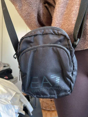 Svart axelväska från Emporio Armani - Snygg svart axelväska från Emporio Armani EA7 med justerbar rem och två dragkedjefack. Väskan är i slitstark textil och har en stilren logga framtill. Perfekt storlek för det viktigaste och passar till många outfits.