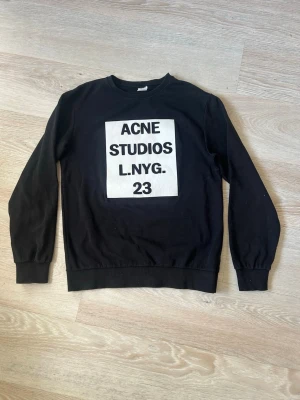 Svart sweatshirt Acne Studios - Svart sweatshirt från Acne Studios med stor vit patch och svart text på bröstet. Tröjan har rund halsringning, långa ärmar och ribbade muddar vid ärmslut och nederkant. Perfekt för en avslappnad och trendig stil.
