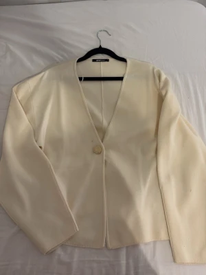 Krämvit cardigan från Gina Tricot - Stilren krämvit tröja från Gina Tricot med v-ringning och en dekorativ knapp framtill. Tröjan har lång ärm och är gjord i ett mjukt, tunt stickat material som känns skönt mot huden. Perfekt att slänga över en topp för en clean look.