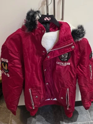 Röd pufferjacka från LEDUNTINO  - Röd pufferjacka från LEDUNTINO med broderade detaljer och patchar på bröstet och ärmen. Jackan har fuskpäls på huvan, dragkedja och knappar framtill samt två fickor med dragkedja. Fodrad med vit teddy på insidan för extra värme.
