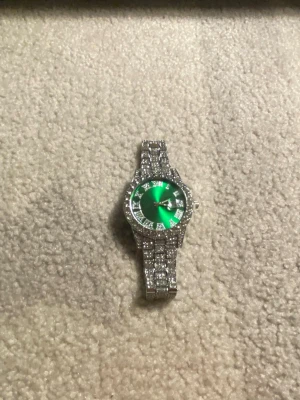 Silverfärgad klocka med grön urtavla - Stilren klocka med glittrande silverfärgat armband och en grön urtavla. Klockan har romerska siffror och är dekorerad med stenar runt boetten och på armbandet. Perfekt accessoar för att sticka ut och ge din outfit extra bling.