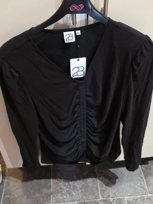 Svart v-ringad blus från 2-Biz - Stilren svart blus från 2-Biz med v-ringning och snygg rynkning framtill. Blusen har långa ärmar och är tillverkad i ett mjukt, lätt material som ger ett elegant fall. Perfekt för dig som gillar en klassisk men trendig look.