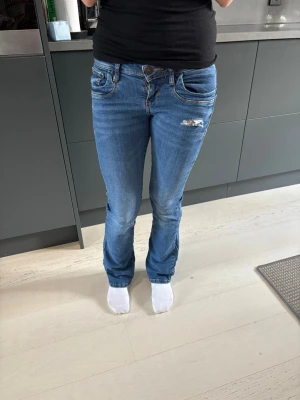 Blå bootcut jeans med slitning - Snygga blå bootcut jeans med klassisk femficksdesign och en cool slitning på höger lår. Jeansen har normal midja och kontrastsömmar. Perfekta för en avslappnad och trendig look.