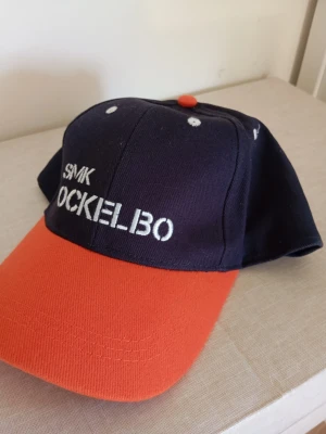 SMK Ockelbo keps i blått och orange - Snygg keps med mörkblå krona och orange skärm, broderad text 'SMK OCKELBO' framtill. Kepsen har vit brodyr och en orange knapp på toppen. Perfekt accessoar för dig som gillar sportig stil.