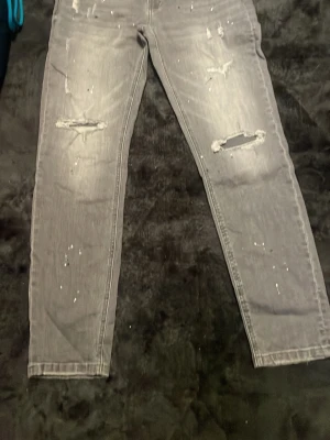 Grå slitna jeans med hål - Säljer ett par grå jeans med slitningar och hål på benen samt stänkdetaljer framtill. Jeansen har rak passform och klassisk femficksdesign. Perfekta för en avslappnad och trendig look.