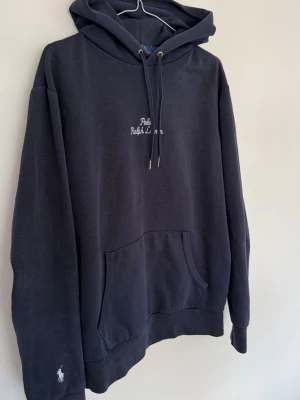 Mörkblå hoodie från Polo Ralph Lauren - Snygg mörkblå hoodie från Polo Ralph Lauren med broderad logga på bröstet och liten vit hästlogga på ärmen. Tyget runt fickan är lite skadat, ingen märkvärt.