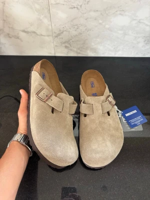 Birkenstock boston - Säljer ett par snygga beige tofflor från Birkenstock i mocka. Modellen har en klassisk design med justerbart spänne i metall och mjuk innersula. Perfekta för dig som gillar stilrena och bekväma skor. Passar både hemma och ute.