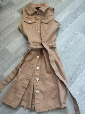 Beige ärmlös klänning med bälte - Snygg beige ärmlös klänning från Madlady i jeansmaterial. Klänningen har knappar framtill, krage, två bröstfickor med lock och ett matchande bälte i midjan. Rå kant nertill för en trendig look. Perfekt för dig som gillar en avslappnad men stilsäker stil.