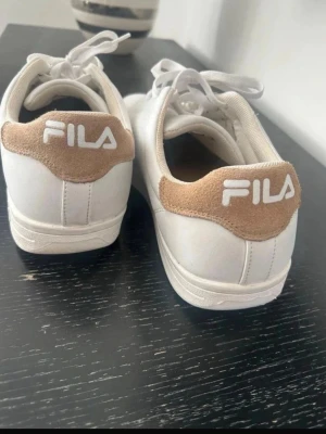 Vita FILA sneakers med beige detaljer - Stilrena vita sneakers från FILA med beige mockadetalj och broderad logga på hälen. Skorna har klassisk låg profil, vita snören och ovandel i syntetmaterial. Perfekta för en clean och sportig look.