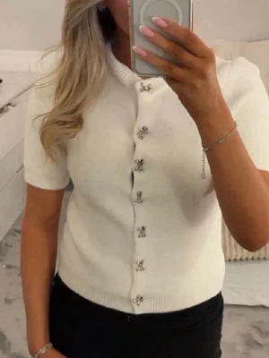 Vit kortärmad kofta med pärlknappar - Söt vit kofta med korta ärmar och dekorativa pärlknappar framtill. Ribbstickad nederkant ger en snygg passform. Perfekt att styla med både jeans och kjol för en fräsch look. M men skulle säga mer som S