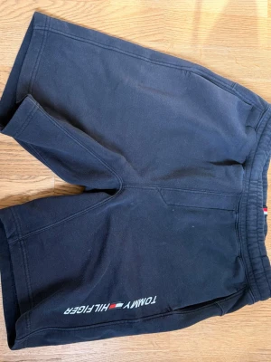 Mörkblå shorts från Tommy Hilfiger - Snygga svarta shorts från Tommy Hilfiger med logga tryckt längs ena benet. De har elastisk midja och är tillverkade i mjukt bomullsmaterial, perfekta för en avslappnad stil.