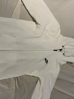 Vit Nike hoodie med dragkedja - Snygg vit hoodie från Nike med dragkedja framtill och svart Nike-logga på bröstet. Utrustad med huva och dragsko samt två fickor på sidorna. Perfekt för en sportig och avslappnad stil.