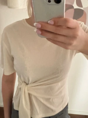 Beige topp med knyt från H&M - Trendig beige topp från H&M med kort ärm och snygg knytning i midjan. T-shirten har rund halsringning och är gjord i mjuk bomull, perfekt för en clean och stilren look.