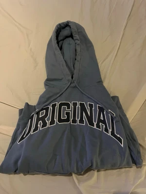 Blå hoodie med tryck 'ORIGINAL' - Snygg blå hoodie med stor vit och svart text 'ORIGINAL' över bröstet. Hoodien har huva med dragsko och en klassisk magficka framtill. Perfekt för en avslappnad och cool stil. Materialet känns mjukt och bekvämt, passar perfekt till jeans eller joggers.