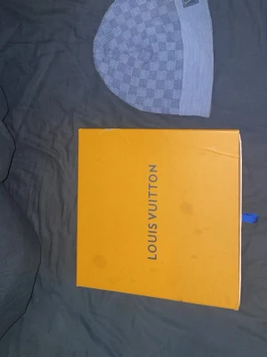 Grå Louis Vuitton mössa - Exklusiv grå mössa från Louis Vuitton med klassiskt rutmönster och diskret logotyp på etiketten. Mössan är stickad och har en mjuk, bekväm passform. Perfekt för dig som vill ha en lyxig accessoar till hösten och vintern.