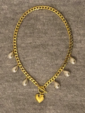 Guldhalsband med pärldroppar och hjärta - Snyggt halsband i guldfärgad rostfritt med fem droppformade pärlor och ett hjärtformat hänge framtill. Kedjan har en chunky design och stängs med en dekorativ T-lås. Perfekt för dig som vill ha ett statement-smycke med trendiga detaljer.