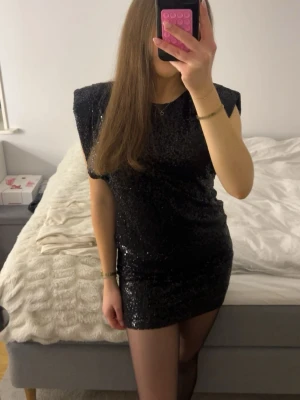 Svart glittrig miniklänning - Snygg svart miniklänning med glittrigt tyg och markerade axlar från Nelly. Klänningen har korta ärmar och en figurnära passform som ger en trendig look. Klänningen är endast använd vid ett tillfälle och är därmed som ny.