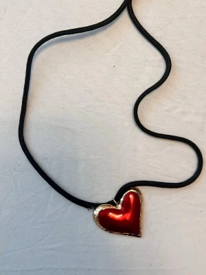 Halsband med rött hjärthänge - Svart halsband med ett stort hjärthänge i röd metallisk färg och guldfärgad kant. Hängsmycket har en tydlig glansig yta och halsbandet har en enkel, stilren design som sticker ut.