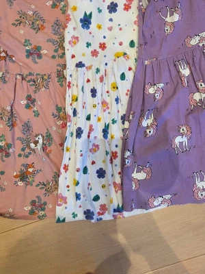 Tre färgglada klänningar med mönster - Tre söta klänningar i bomull: en rosa med blommor och djur, en vit med färgglada blommor och fjärilar, samt en lila med enhörningar. Alla är ärmlösa och har en kort klänningslängd. Perfekta för sommarens varma dagar.